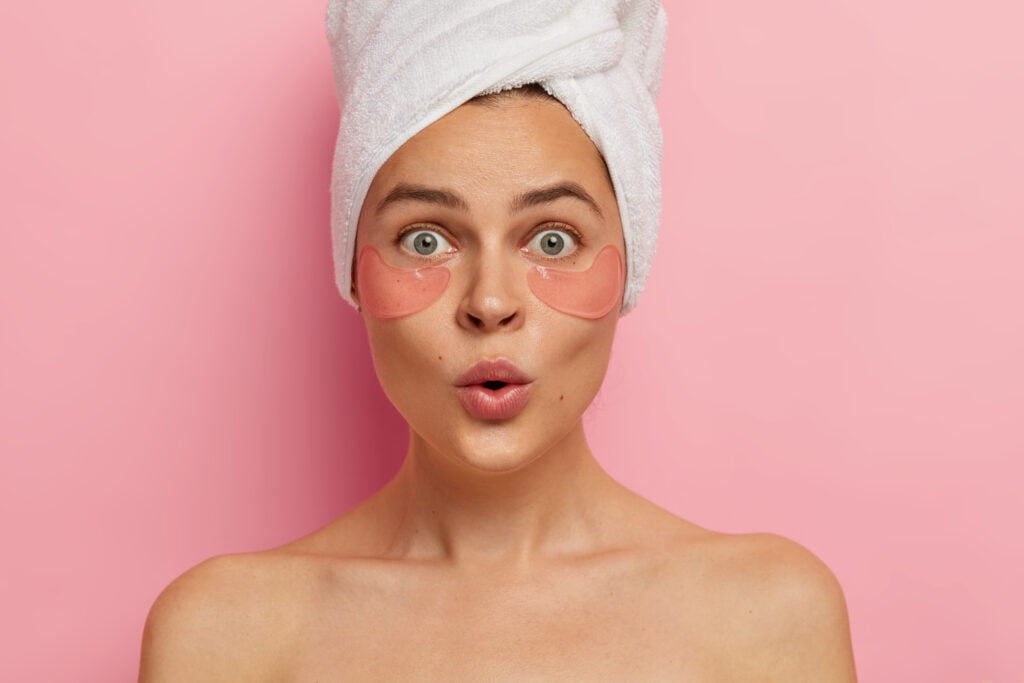 Under eye treatment and body care concept. Shocked young Caucasian woman applies beauty patches, removes dark circles and puffiness, looks surprisingly at herself in mirror, stands topless indoor
